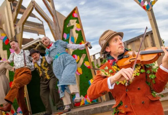 Musician playing violin at outdoor event with colourful decorated wooden structures and visitors in background.