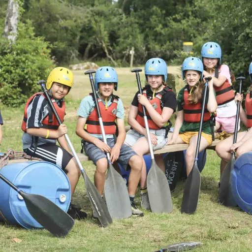 Children in helmets and life jackets sitting with paddles before a water sports activity.
