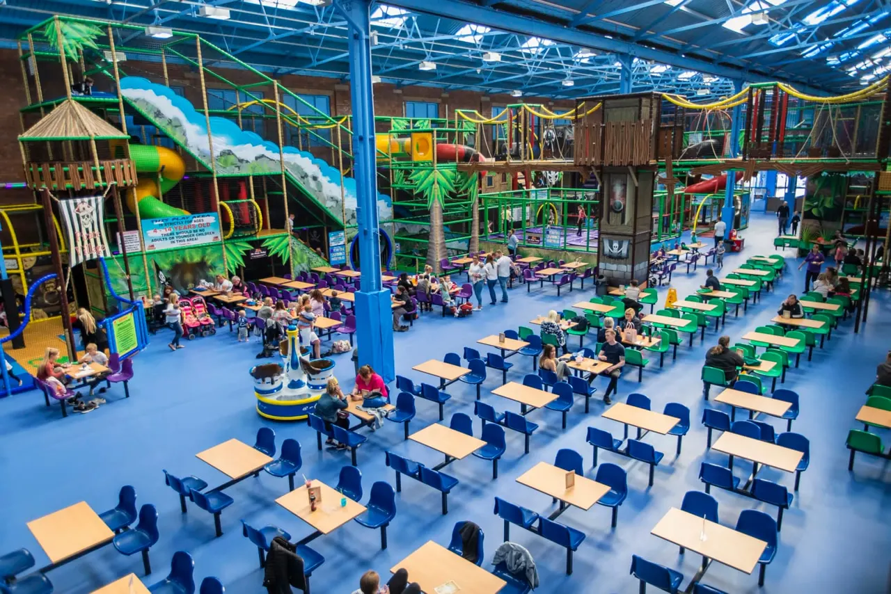 Interior of an indoor play centre with children and families dining at picnic tables, with colourful climbing structures and slides visible in the background.