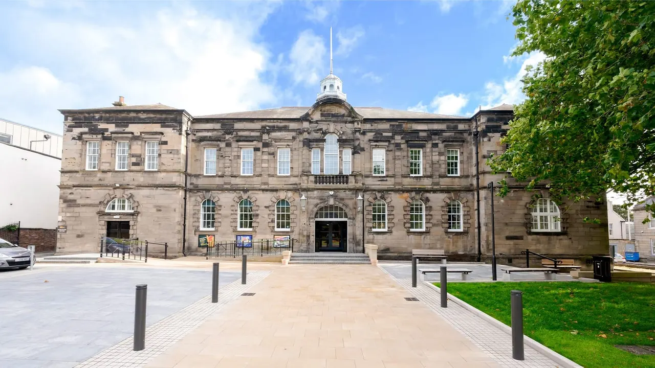 A grand stone theatre building with a spire, viewed from its paved forecourt on a sunny day.