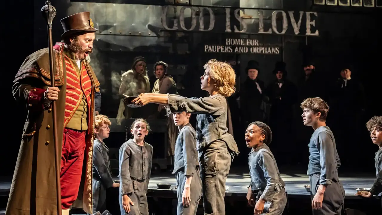 Actor in ornate period costume addresses group of children in ragged clothes on theatre stage.