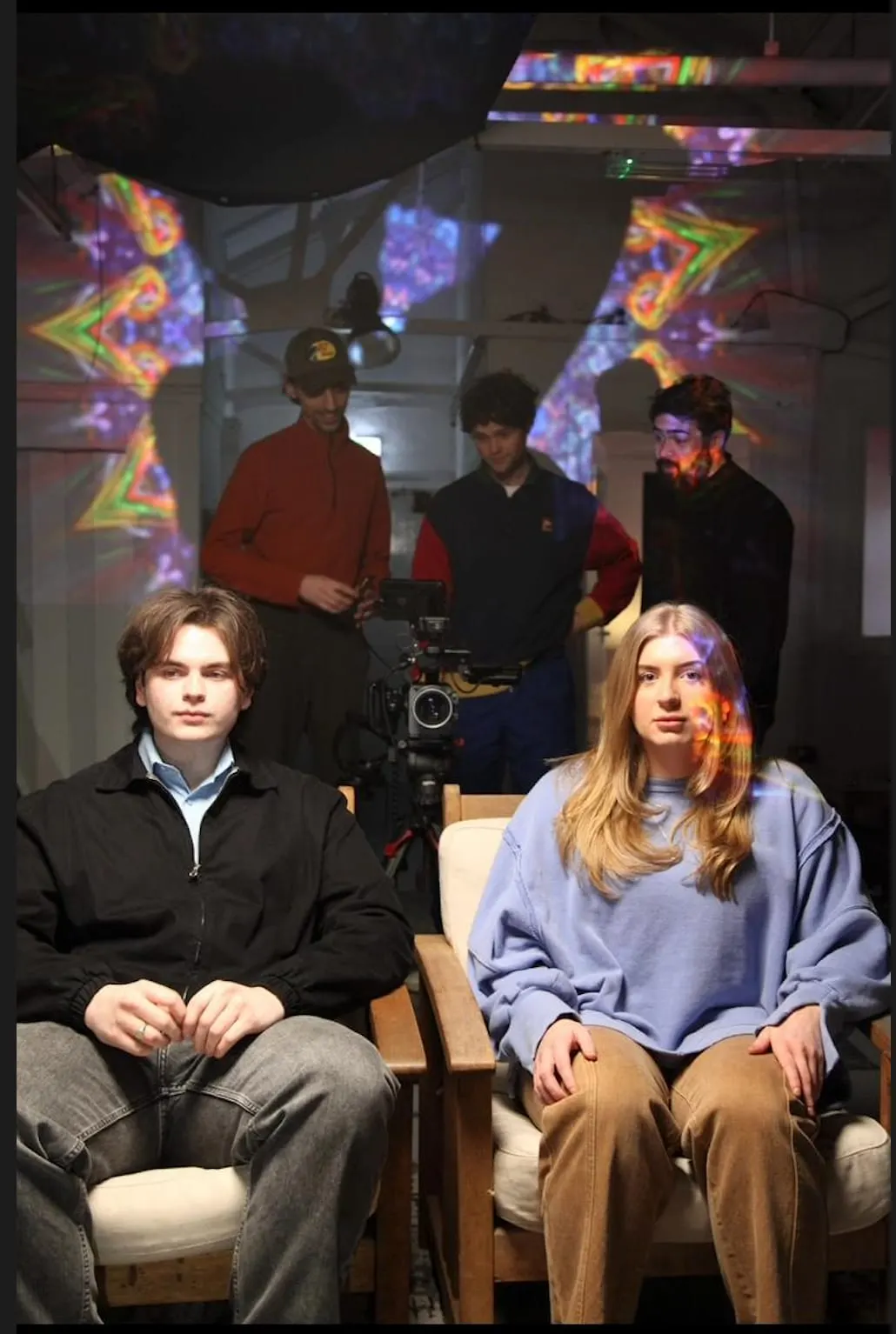 A young man and woman sit in chairs facing the camera whilst others stand behind them near film equipment, surrounded by colourful projected light patterns.
