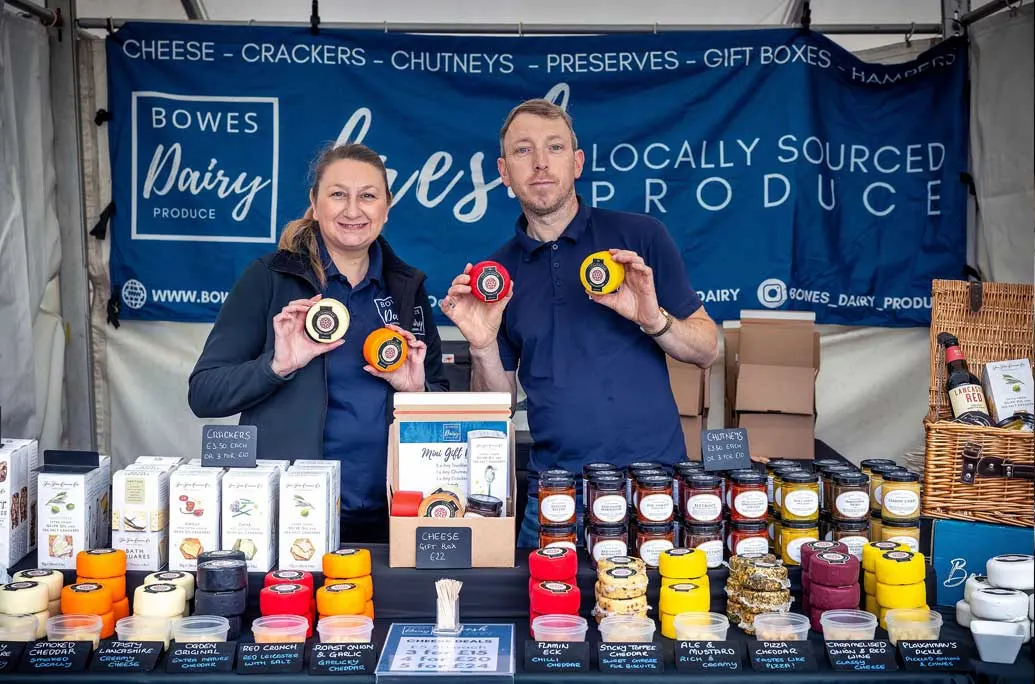 Two people at a farm shop stall displaying locally-sourced dairy products, cheeses, chutneys and preserves.