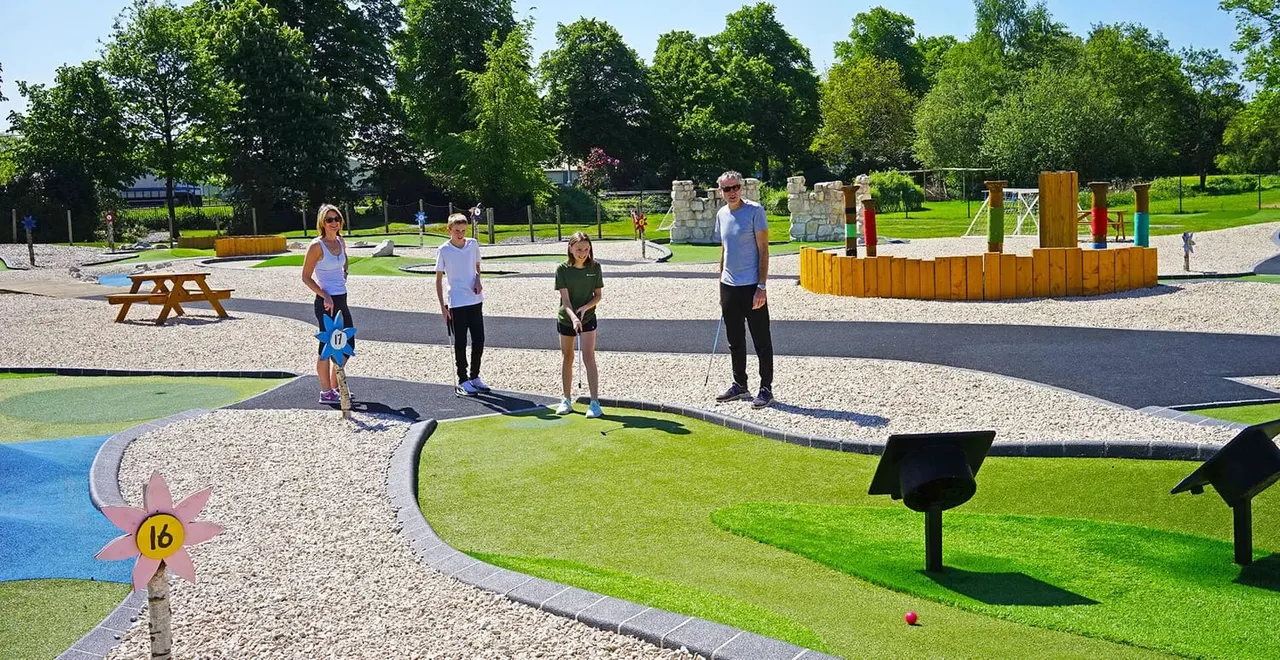 Family and friends playing mini golf on a sunny day at an outdoor course with trees and play equipment in the background.