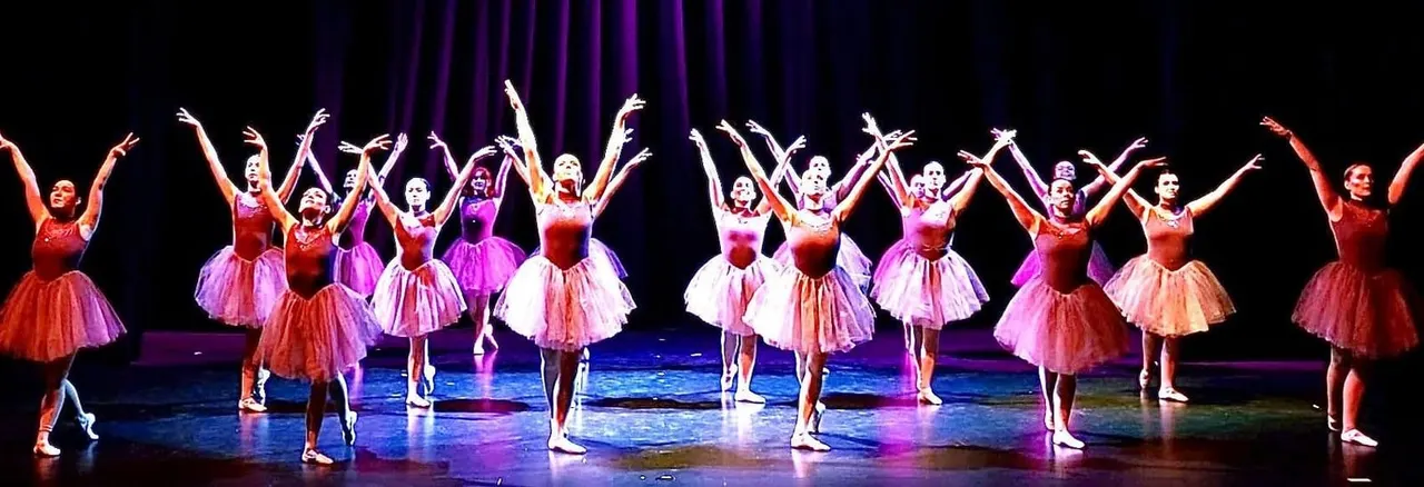 A line of young ballet dancers in pink and red tutus perform on stage with arms raised, lit by purple and blue stage lighting.