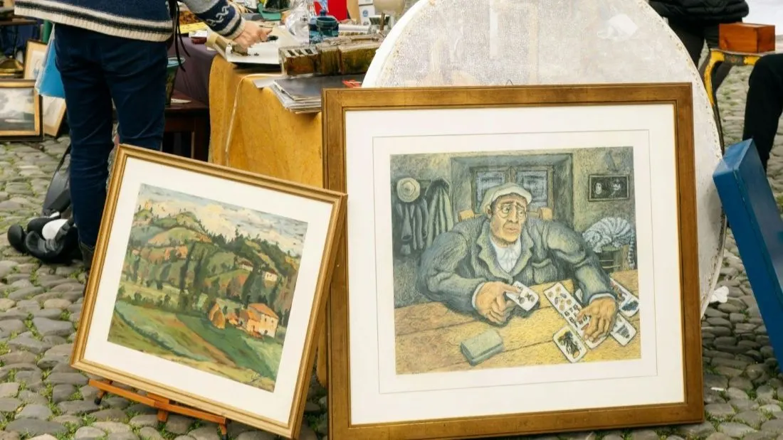 Framed artworks displayed at an antiques or art market stall, with visitors browsing in the background.