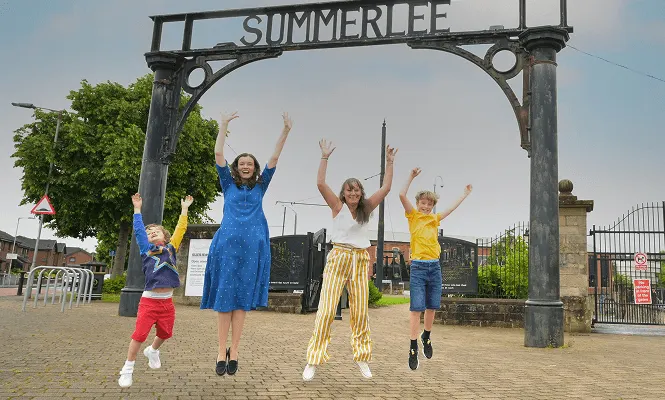 Four children jumping with arms raised beneath a heritage iron gateway sign at an outdoor museum venue.