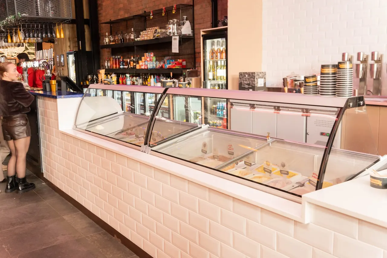 Ice cream parlour counter with glass display cases showing flavours, customer approaching, brick walls and shelves of bottles behind.