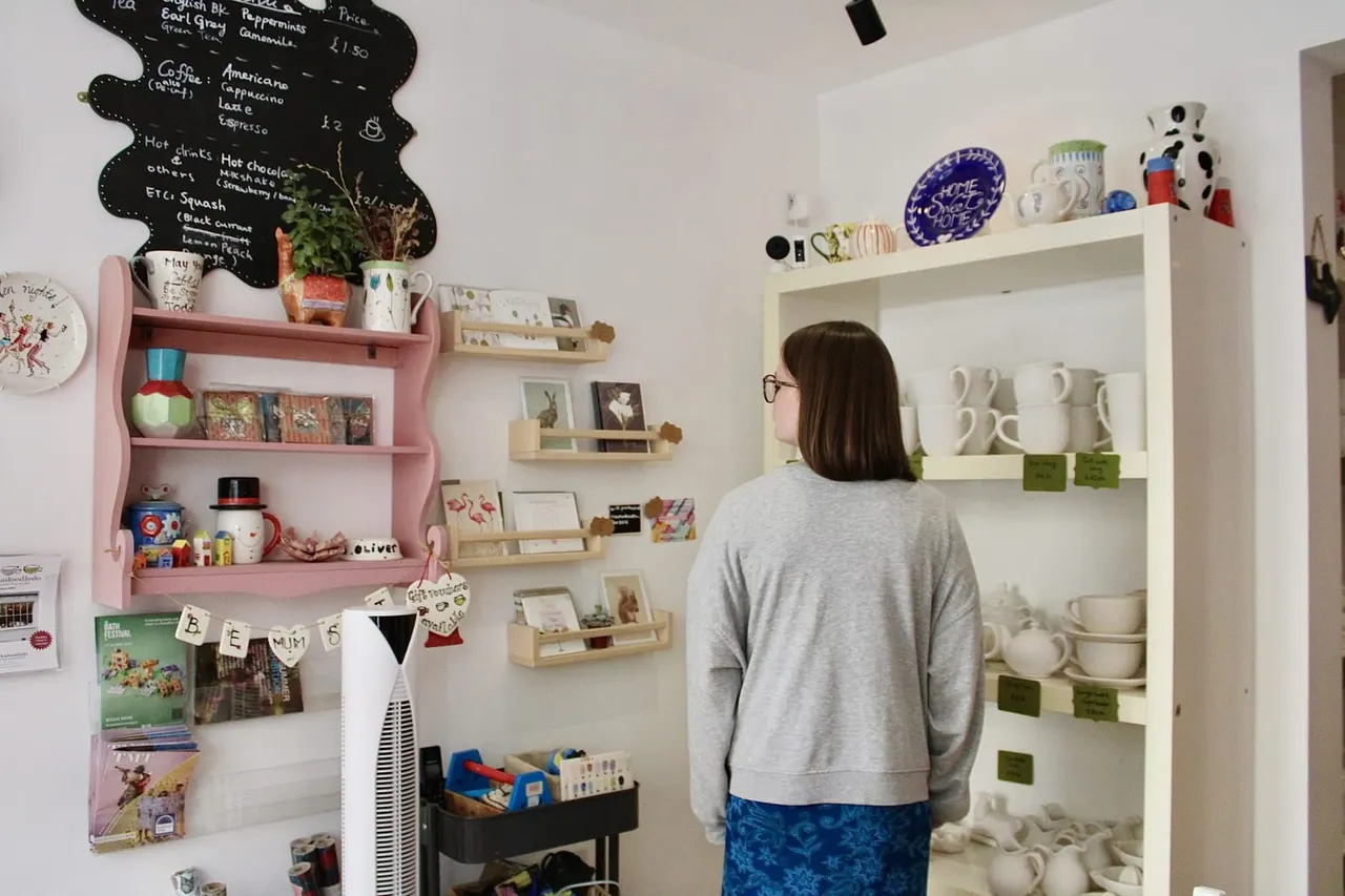 A person wearing glasses and a grey sweatshirt views shelves displaying ceramics, mugs, and decorative items in a bright, colourful café interior.