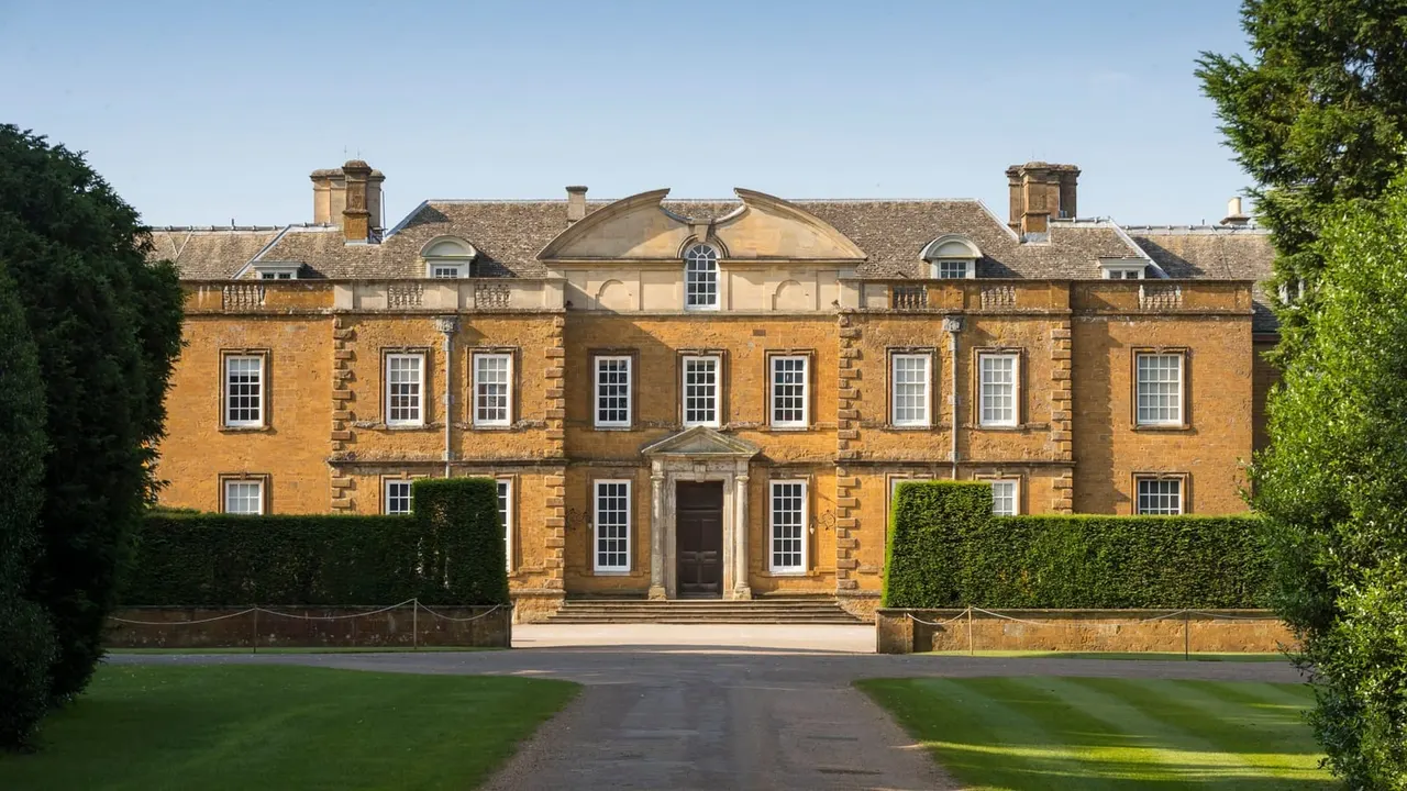 Honey-coloured stone country house with manicured gardens and driveway on a clear day.