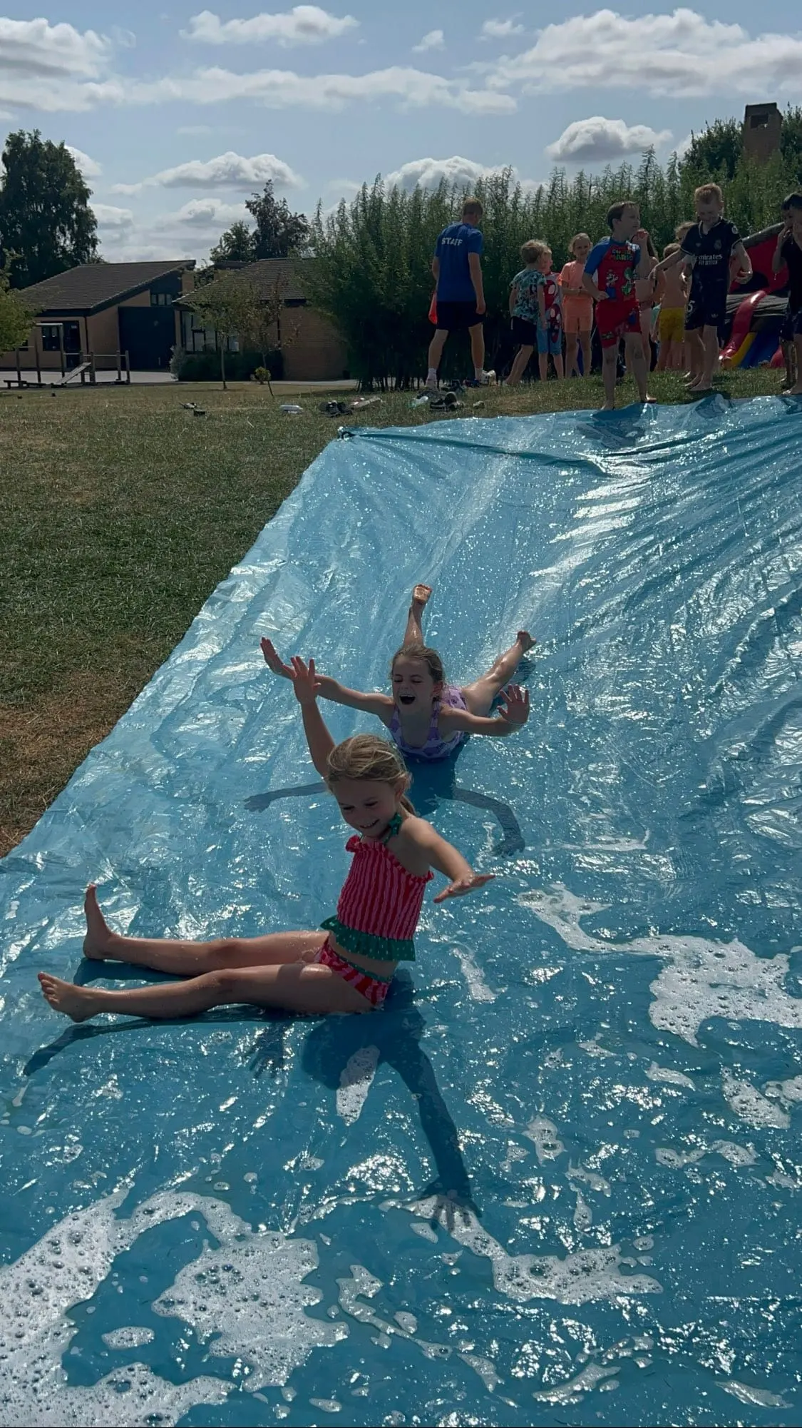 Children sliding down a wet blue slip-n-slide on grass, with other children waiting in line nearby and a building behind.