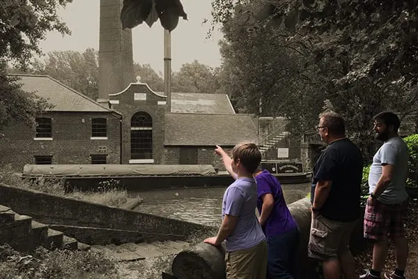 Visitors stand by a canal lock watching a brick industrial building with a tall chimney across the water.