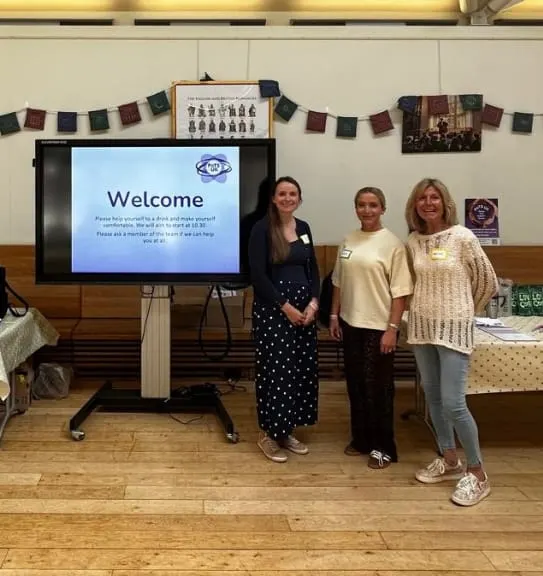 Three women stand beside a screen displaying a welcome message at an indoor event venue.