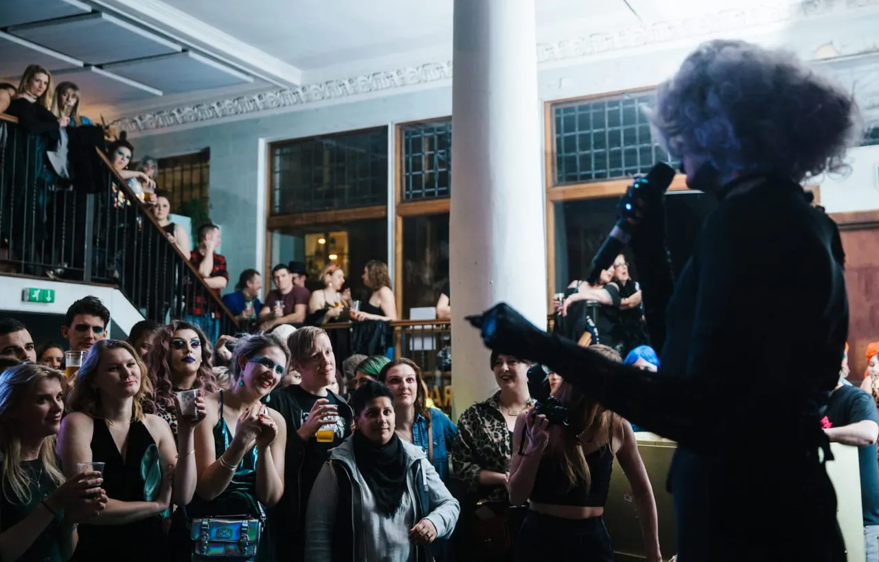 A performer with blue-tinted hair sings into a microphone to a crowd of spectators in a multi-level indoor venue space.