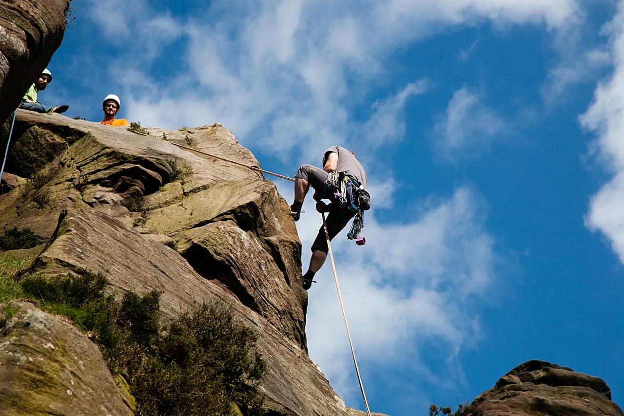 Person rock climbing on outdoor stone face with rope and safety equipment, whilst instructors observe from the cliff top.