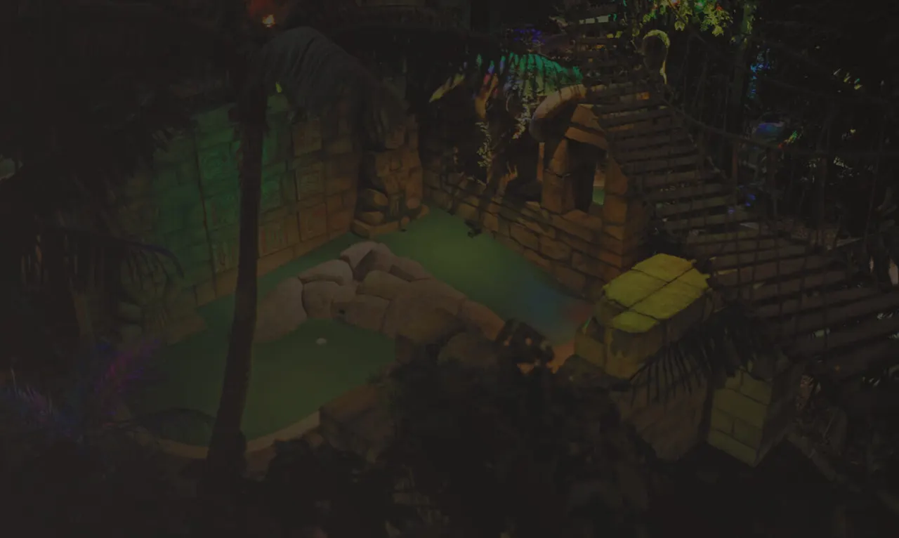 Children playing mini golf in an indoor course with green flooring and wooden structures lit with coloured lighting.