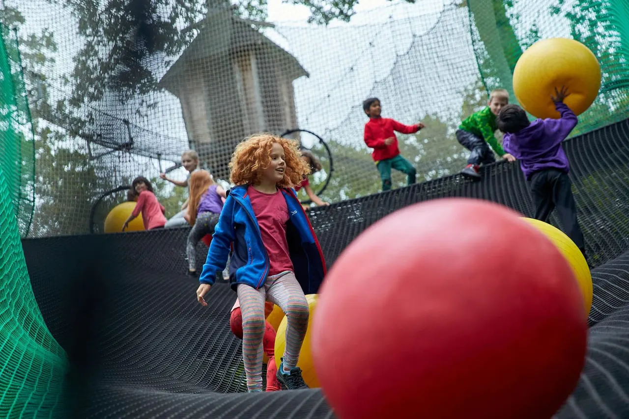 Children bouncing and playing on a trampoline park with large coloured balls and safety netting.