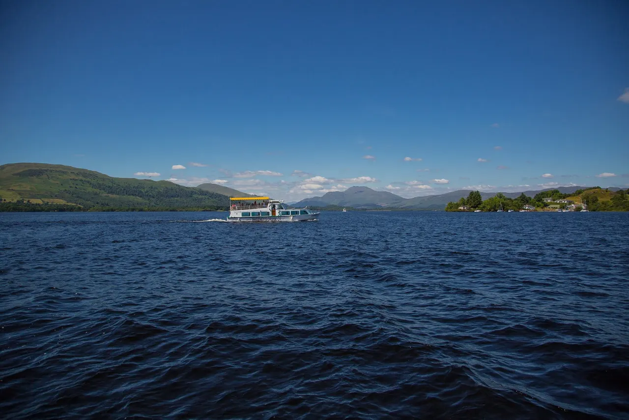 Passenger cruise boat on deep blue loch waters with green mountains and islands in background under clear sky.