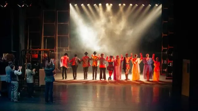 Theatre audience watching performers in red and orange costumes taking a bow on an illuminated stage.