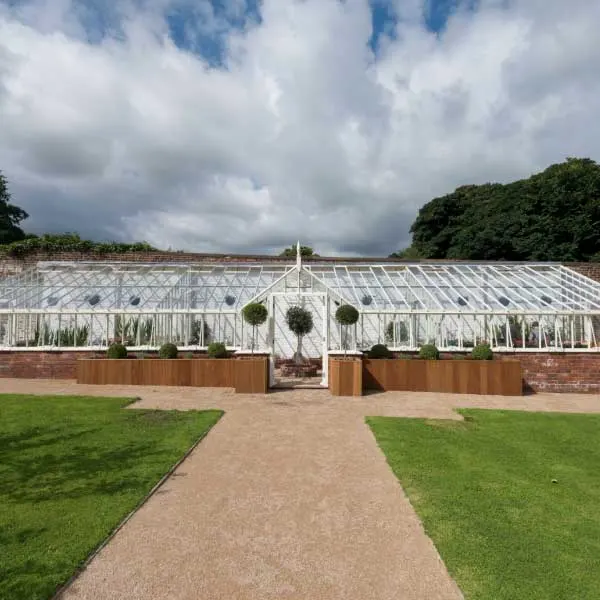A large white Victorian-style greenhouse with manicured lawns and a gravel pathway leading to its entrance under a cloudy sky.