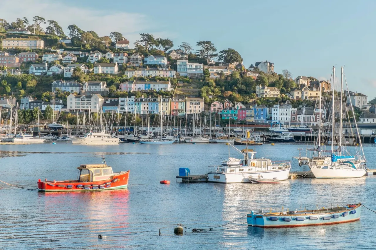 Colourful fishing and sailing boats moored in a calm estuary with a hillside harbour town of pastel-painted houses behind.