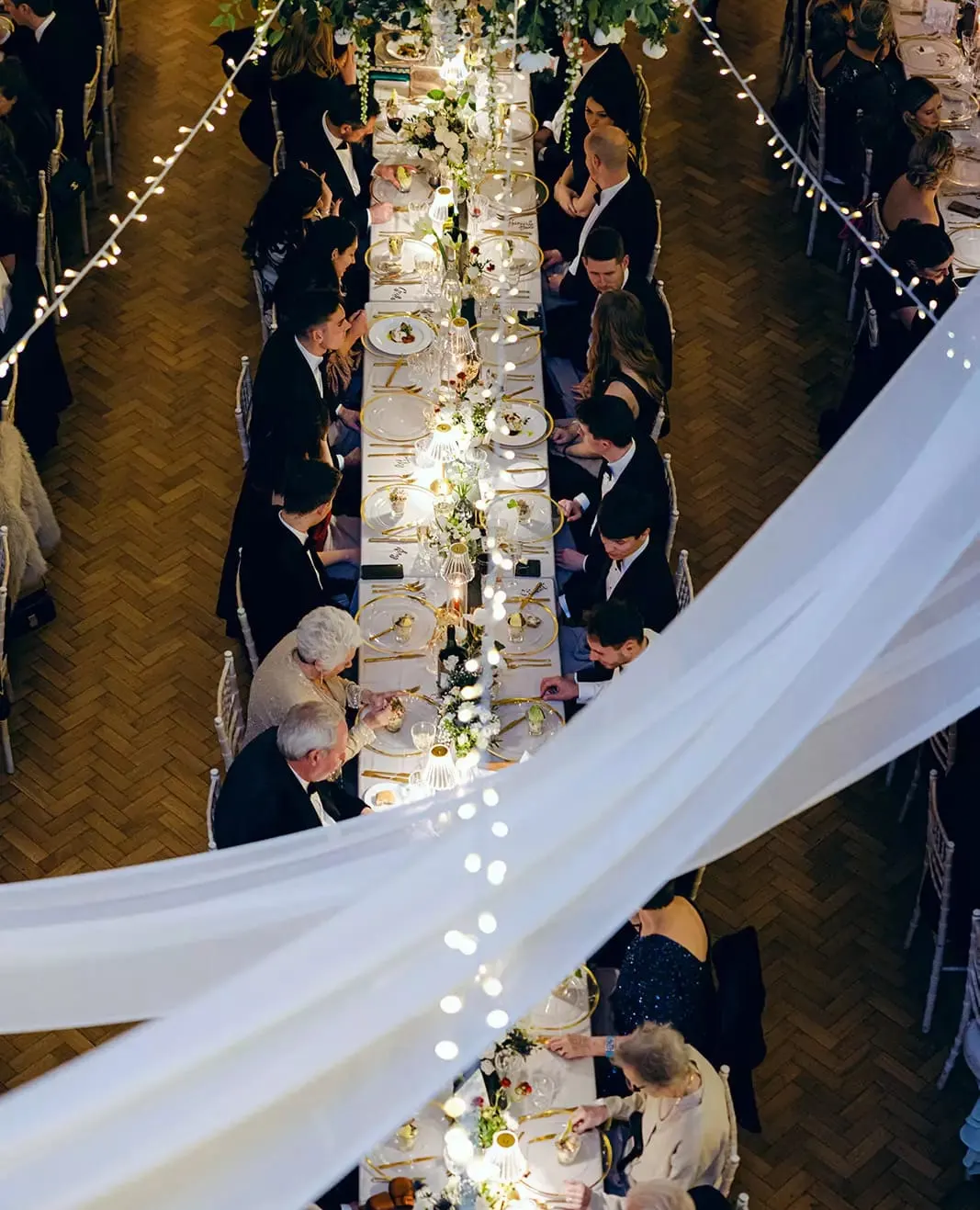 Overhead view of formal dining event with guests in dark attire seated along a long table decorated with greenery and string lights.