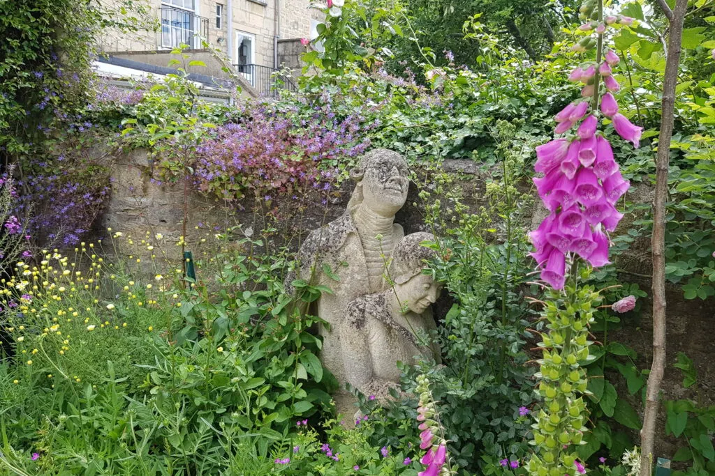 Stone statue surrounded by flowering plants and climbing ivy in a lush garden, with a stone building visible behind.