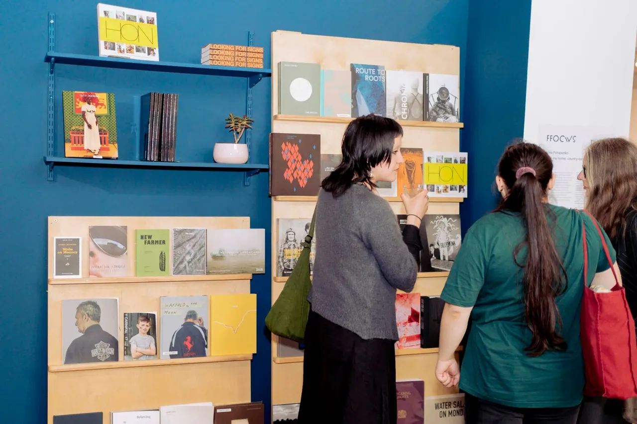Three visitors browse displayed books and artworks on wooden shelves against a bright turquoise wall in a gallery space.