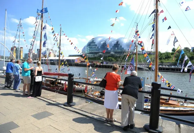 Visitors stand on a waterfront promenade viewing tall ships decorated with colourful flags moored at the marina.