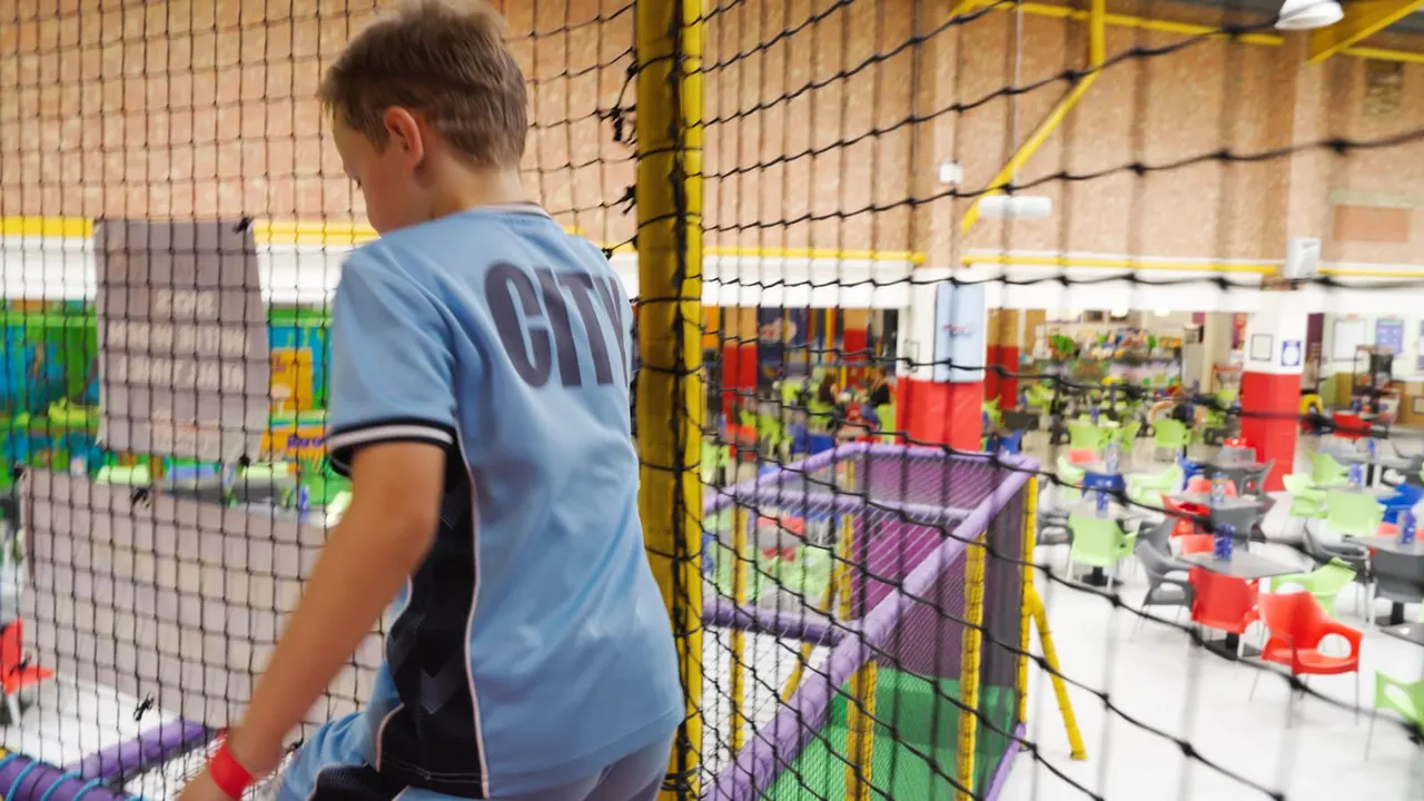Child in blue shirt climbing through netting inside a brightly coloured soft play arena with various play equipment.