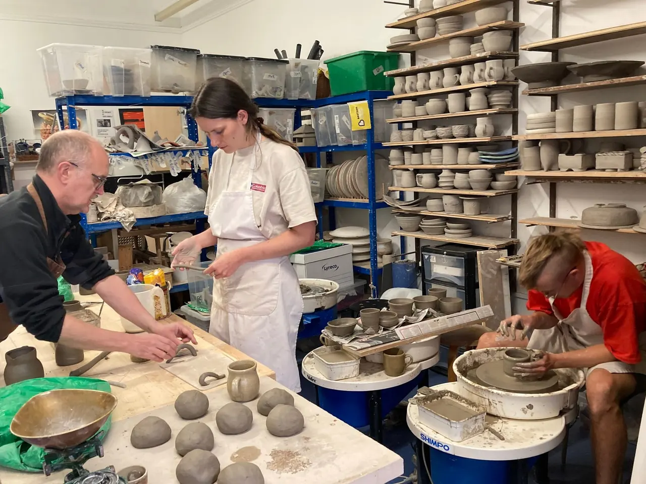 Three people working with clay in a pottery studio, surrounded by shelves of finished ceramic vessels and works in progress.