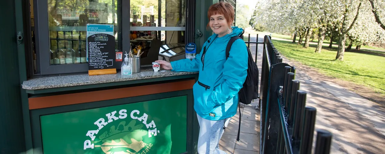 Woman in blue jacket holding a drink at outdoor café counter with blooming trees beyond.