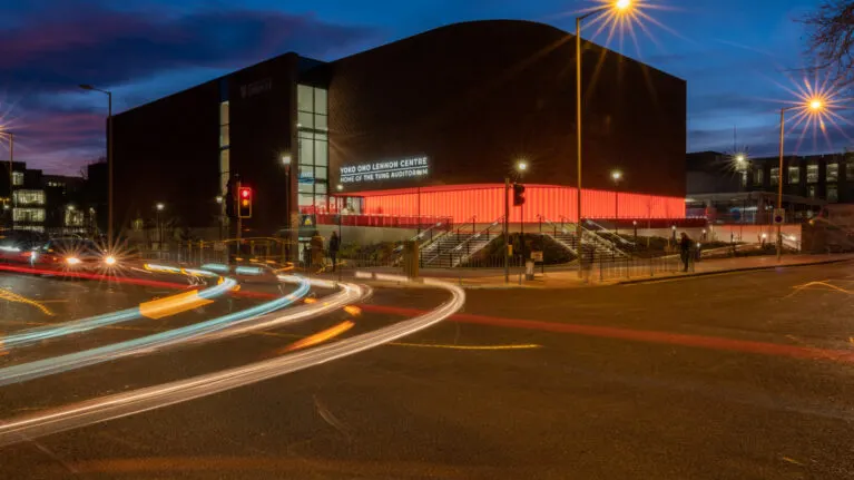 Modern brick building with red entrance canopy lit at dusk, surrounded by street lights and light trails from passing traffic.