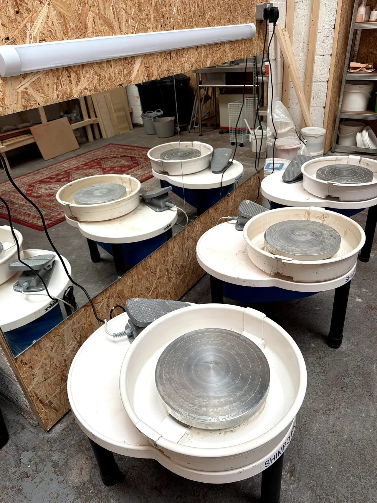 Multiple pottery wheels with grey clay work surfaces arranged in a studio workshop space with OSB walls and shelving.