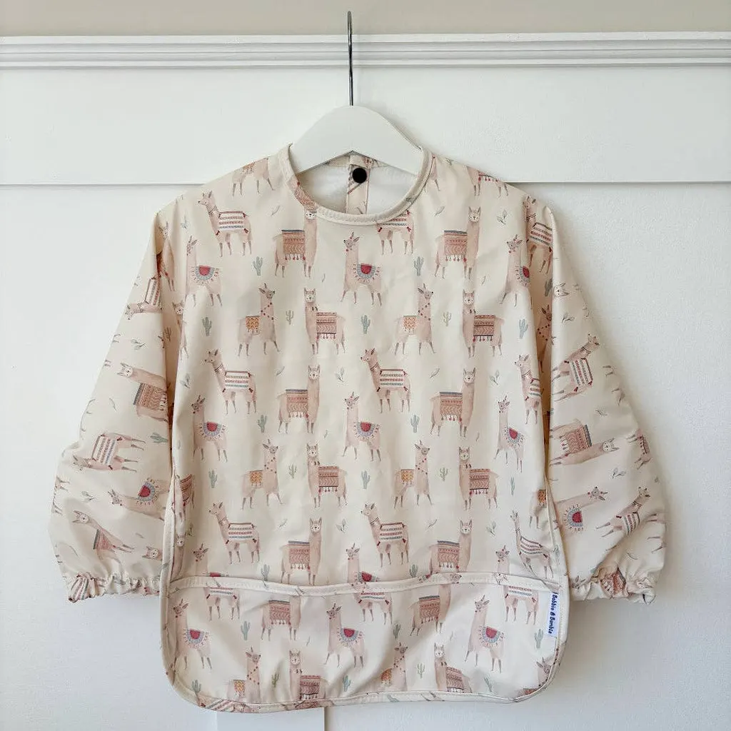 Children's cream sweatshirt with llama and cactus pattern, hung on white hanger against white wall.