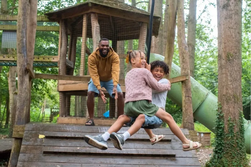 Adult and two children playing on wooden treehouse structure surrounded by tall trees.