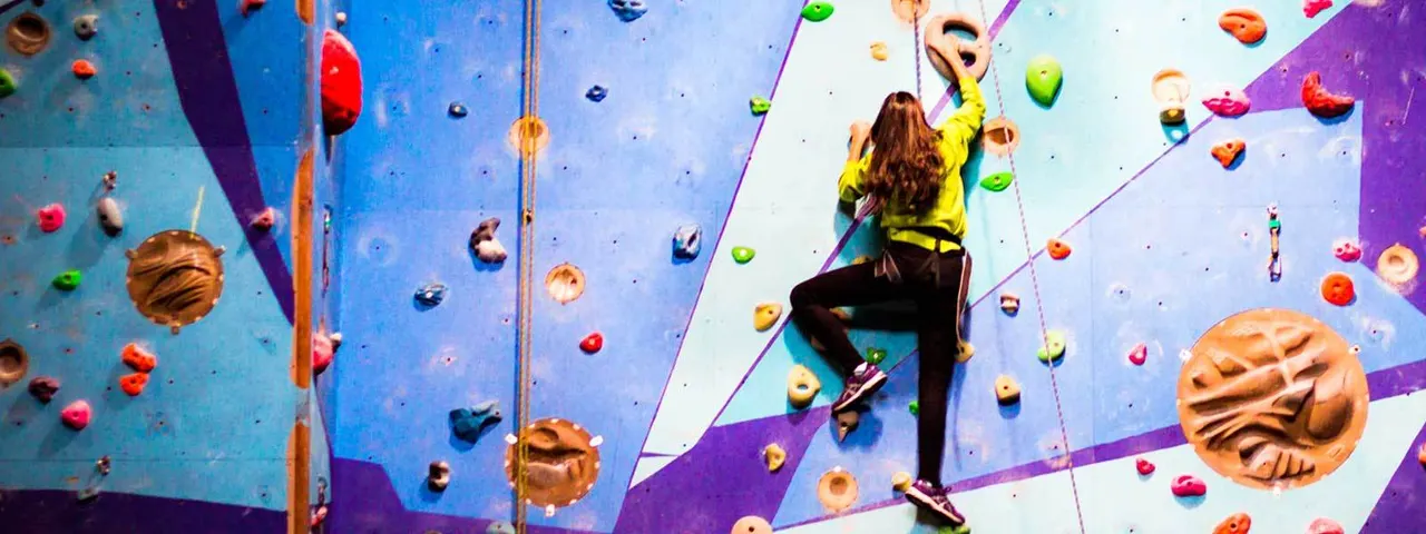 Person in yellow jacket climbing an indoor rock wall with coloured handholds and rope, secured by safety equipment.