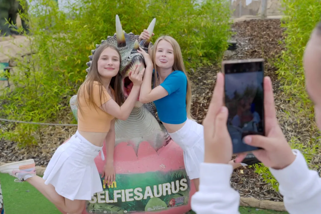 Two young visitors posing with a colourful dinosaur prop whilst someone photographs them on a mobile phone at an adventure golf venue.