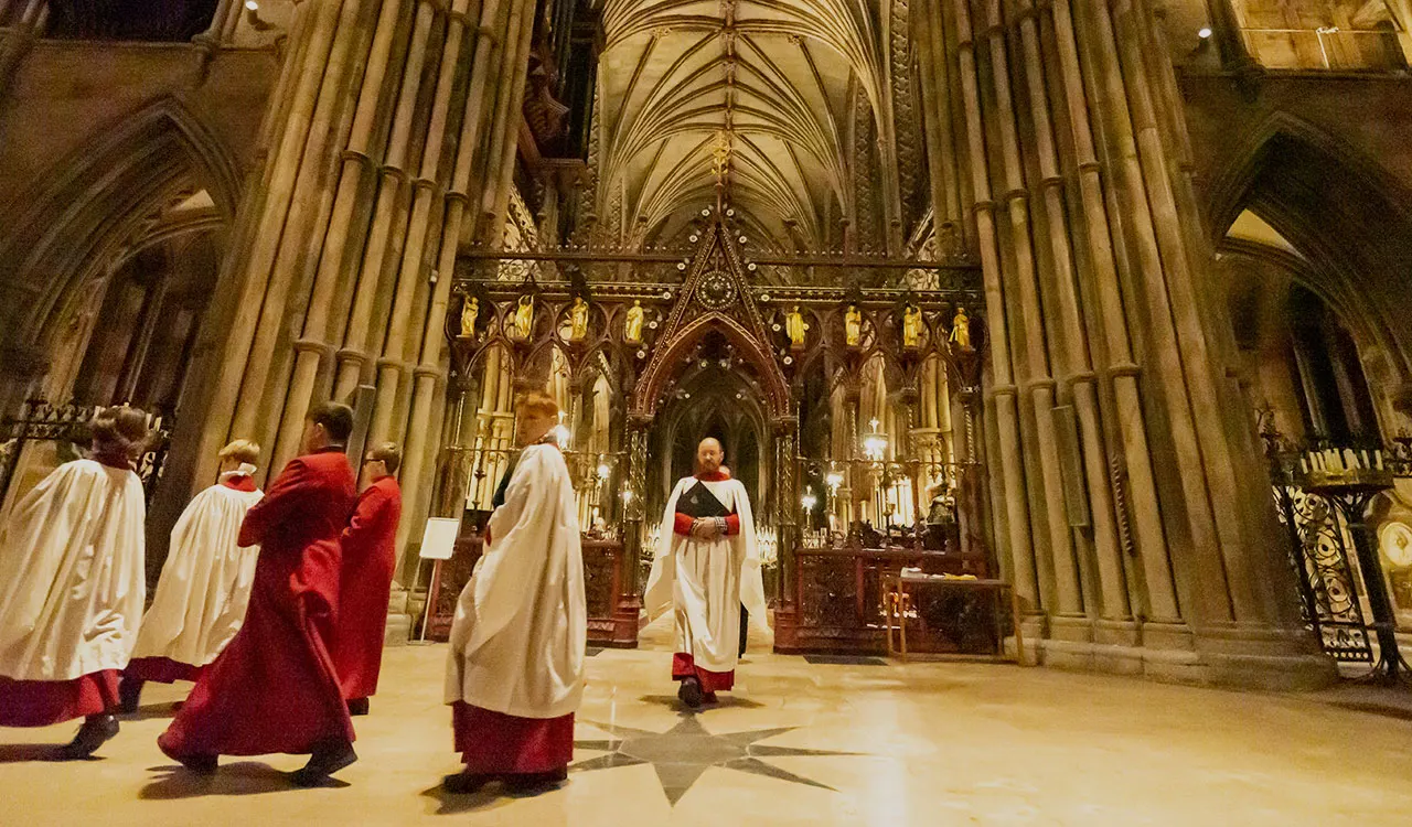 Clergy in red and white vestments walk through the nave of a Gothic cathedral with soaring ribbed vaults and ornate altar.