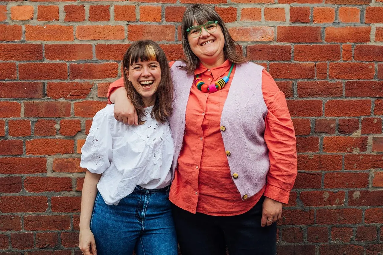 Two smiling women posing together against a red brick wall.