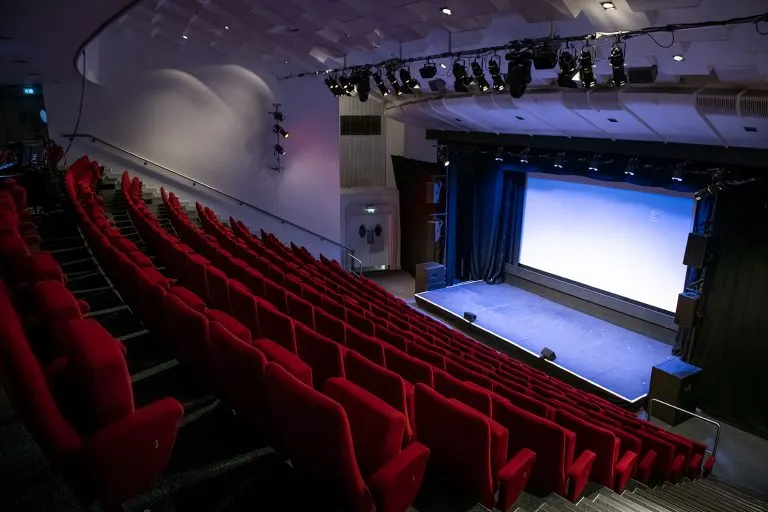 Empty cinema auditorium with rows of red seats facing a screen and stage with professional lighting rigs overhead.