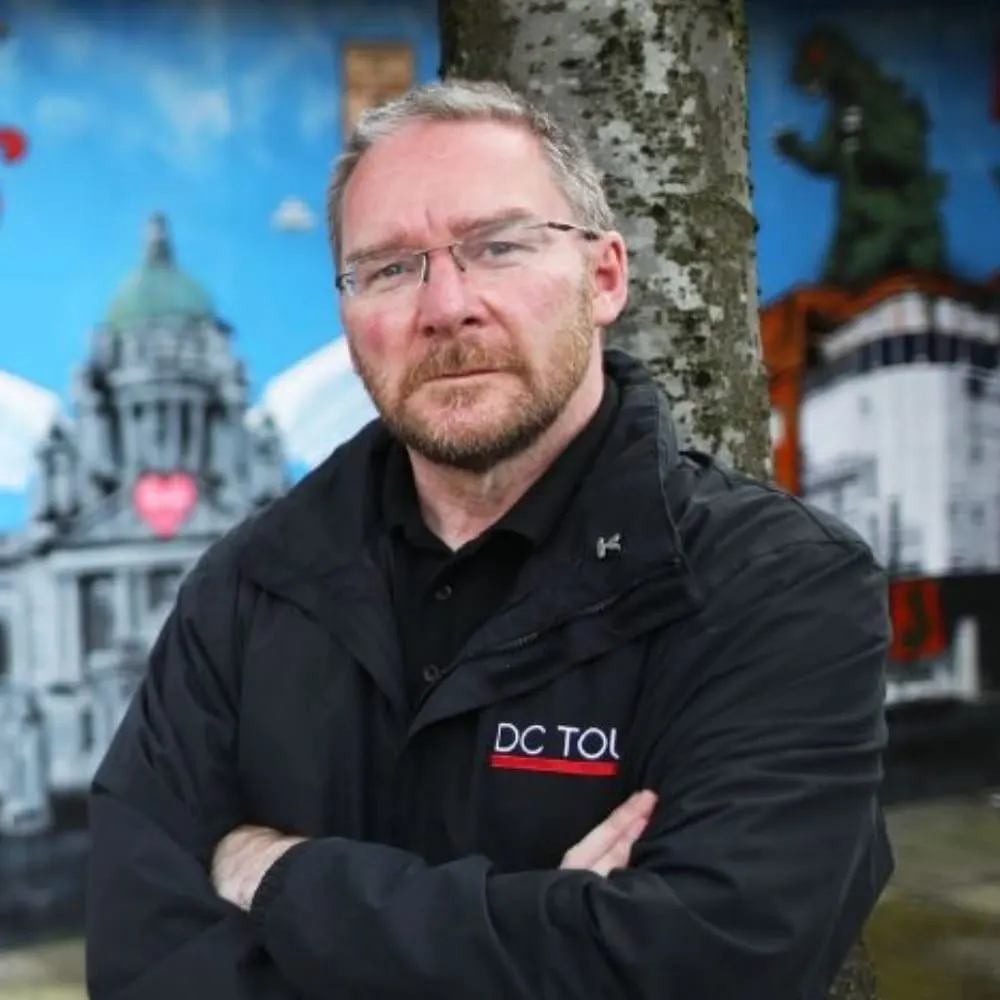 Man wearing glasses and black jacket with arms crossed, standing by a tree with historic buildings in blurred background.