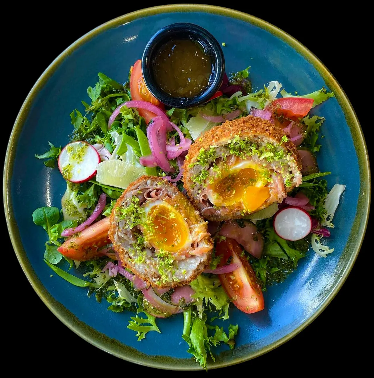 Scotch eggs with pistachio crust on mixed leaves with pickled onions and dressing on blue plate.