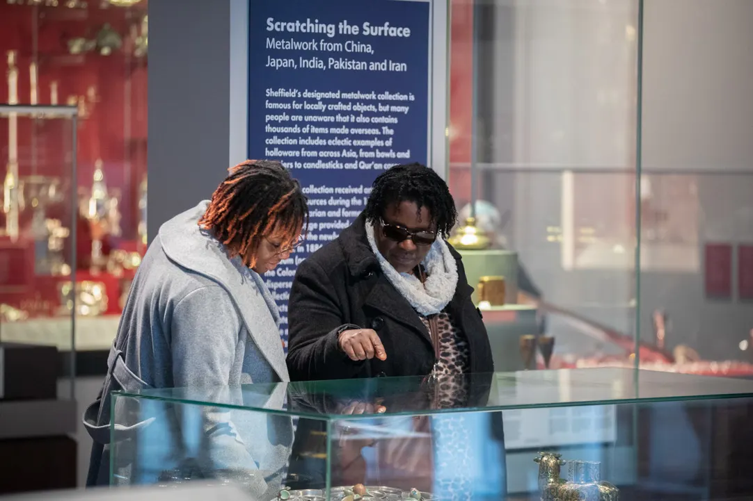 Two visitors viewing a museum display case about metalwork from Asia, with an interpretive panel visible in the background.