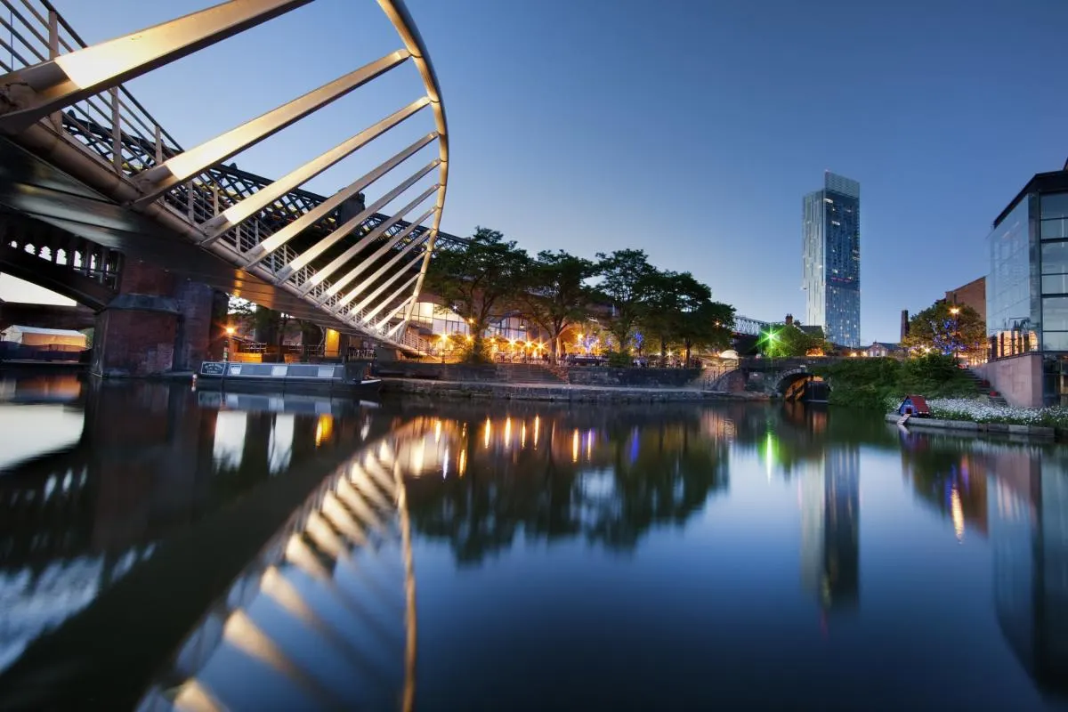 Illuminated footbridge spanning a calm canal at dusk with modern buildings and trees reflected in the water.