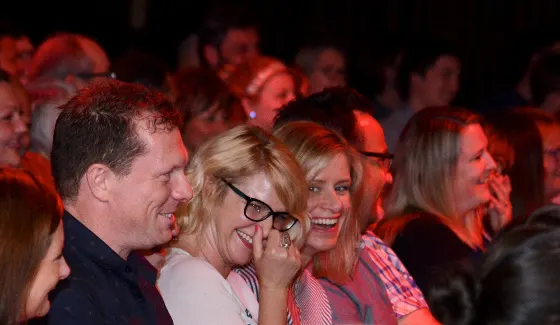 Audience members laughing and smiling whilst watching a comedy performance in a theatre venue lit by red stage lighting.