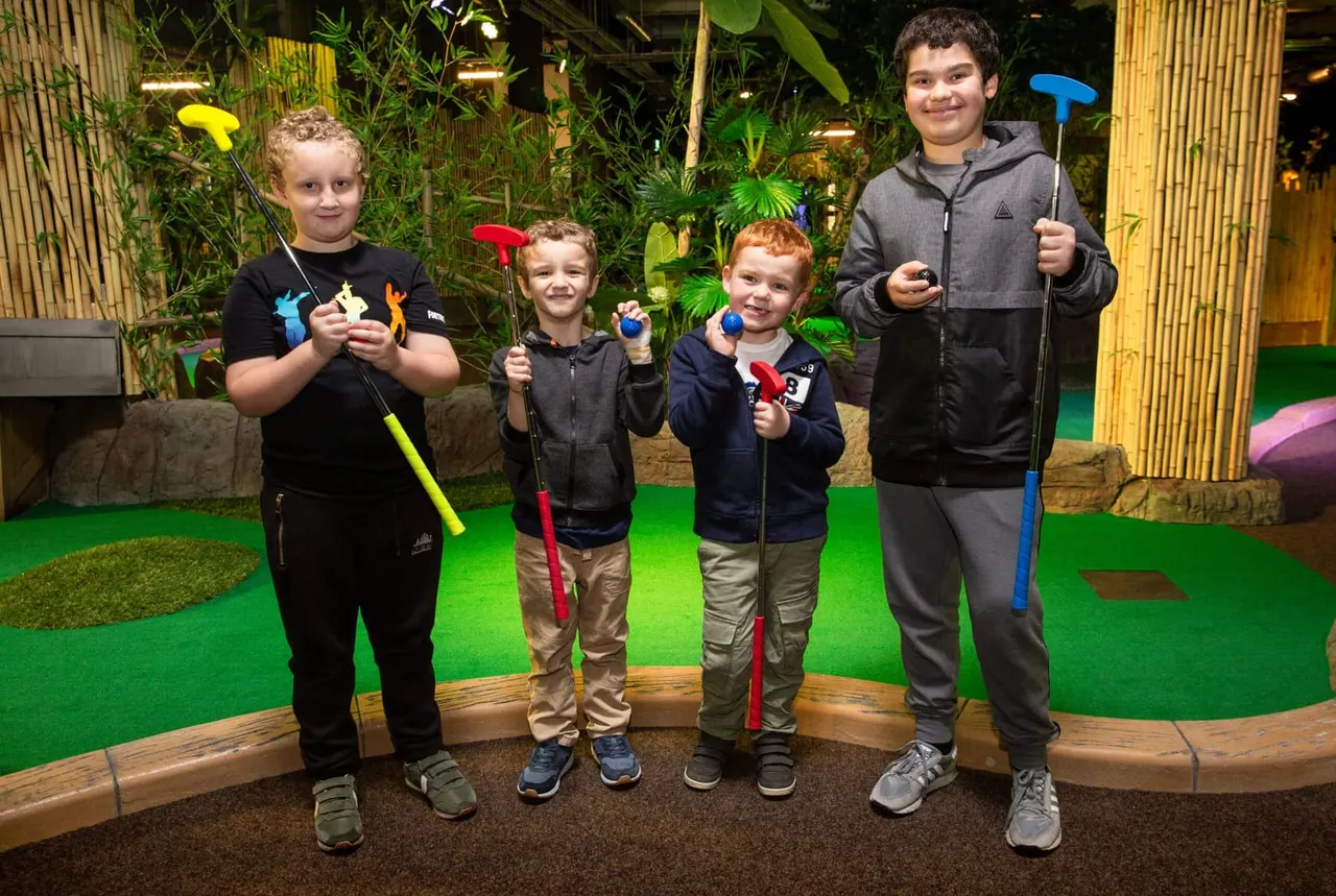 Five children holding coloured putters on an indoor mini golf course with green carpet and tropical plant décor.