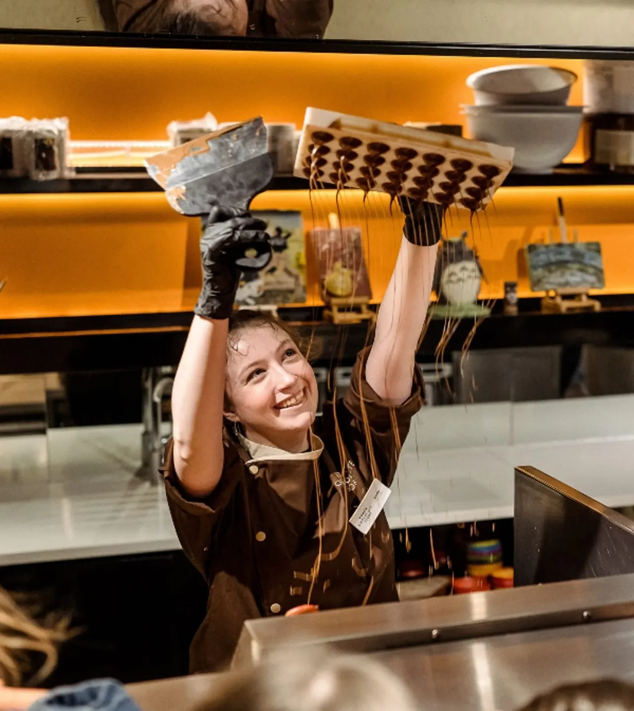 Child in brown uniform holding patterned noodle strainers aloft in a kitchen workshop setting.