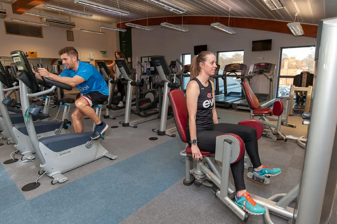 A man and woman exercise on stationary bikes in a gym with various cardio and weight machines.