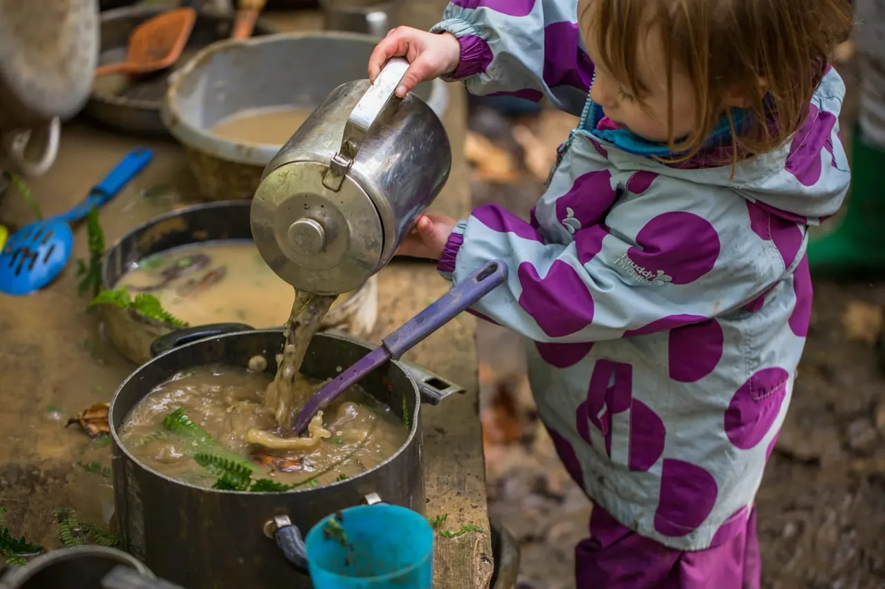 Child pouring water from a metal can into a pot during an outdoor cooking or nature activity session.